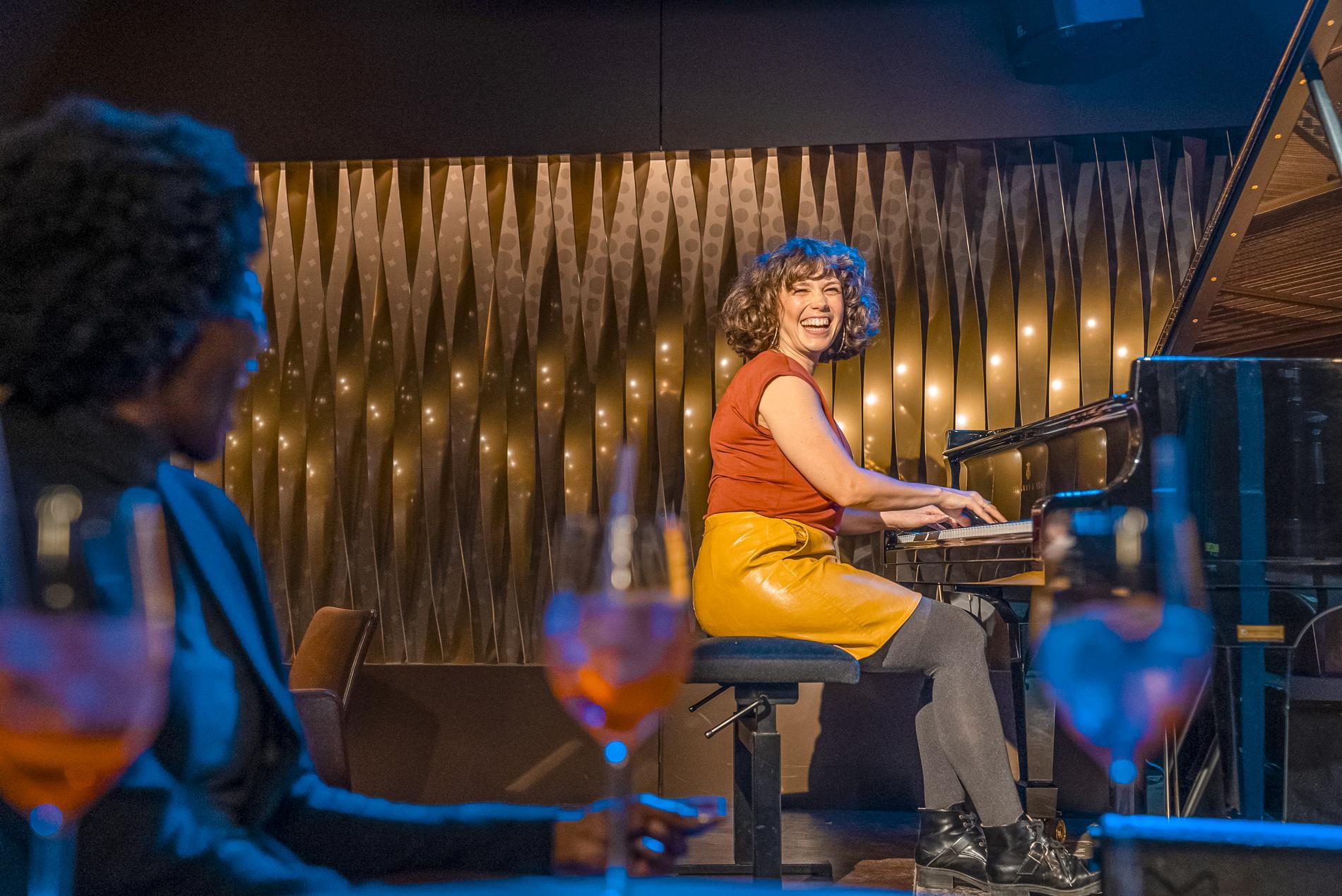 Eine Frau spielt lachend Klavier auf einer Bühne. Ihr Blick ist ins Publikum gerichtet. Im Vordergrund ist unscharf eine weitere Frau und Weingläser erkennbar.